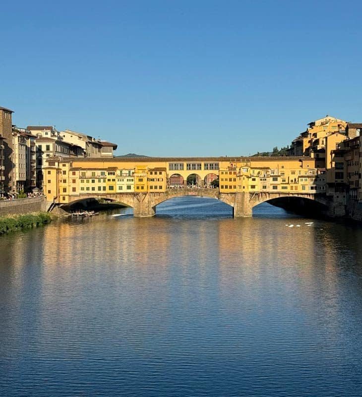 View of the Ponte Vecchio bridge in Florence, Italy, reflecting in the calm waters of the Arno River under a clear blue sky.
